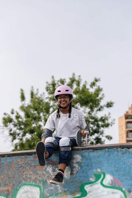 Cuerpo completo de alegre joven afroamericana con camiseta blanca con casco y almohadillas de seguridad con monopatín sentado en la rampa de skate en el parque - foto de stock