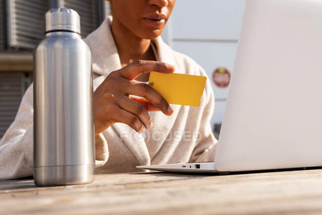 Crop mujer anónima con tarjeta de crédito en la mano escribiendo en netbook moderno al hacer la compra en línea en la calle en la ciudad - foto de stock