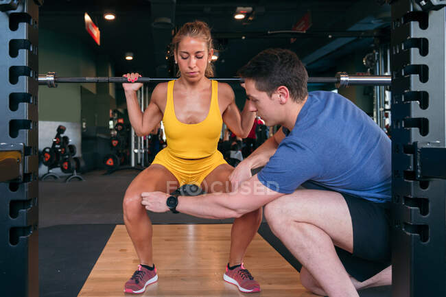 Athlète féminine faisant squats avec haltère lourde pendant l'entraînement dans la salle de gym sous la supervision de l'entraîneur personnel masculin — Photo de stock