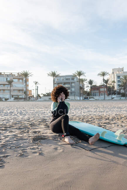 Позитивная женщина с вьющимися волосами в гидрокостюме, разговаривающая по телефону на смартфоне, сидя на песчаном пляже с доской для серфинга — стоковое фото