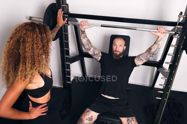 Зверху концентрований спортсмен з татуюваннями підйомний штанга з важкими вагами під час функціональних тренувань виграє жінку-інструктор в тренажерному залі — стокове фото