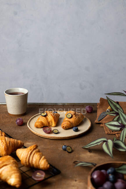 De savoureux croissants frais cuits au four servis dans une assiette avec des fruits placés près d'une tasse de thé sur une table en bois le matin dans une pièce lumineuse — Photo de stock