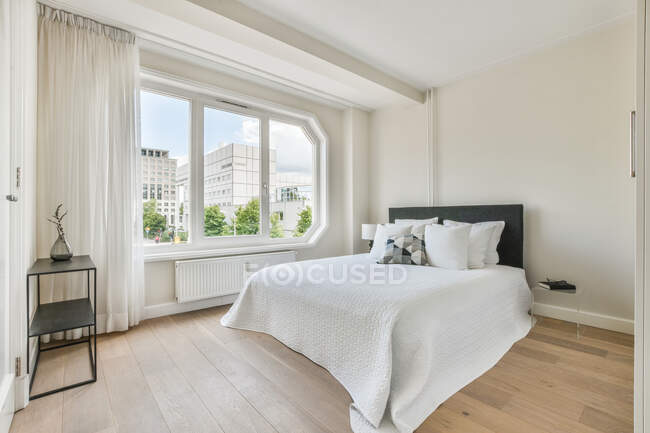 Cómoda cama con manta y almohadas situada cerca de la ventana en el dormitorio soleado de apartamento moderno - foto de stock
