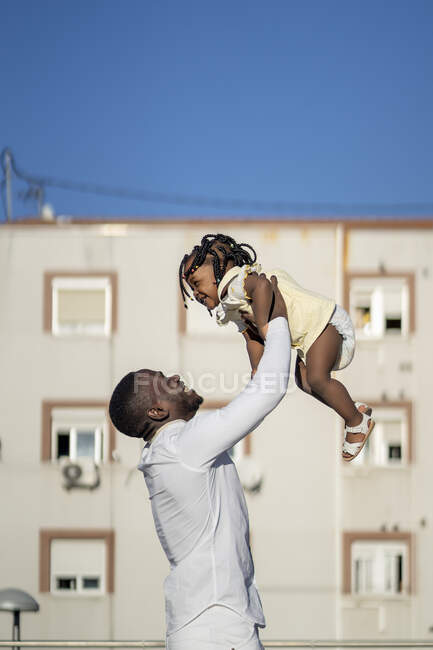 Вид сбоку афроамериканского мужчины, стоящего и поднимающего счастливую маленькую дочь, веселясь на улице в солнечный день — стоковое фото