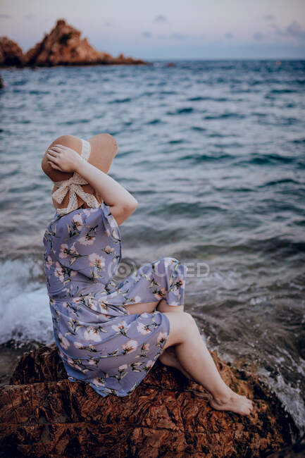 Encantadora joven hembra en vestido de verano y sombrero sentado en la costa rocosa mientras mira hacia otro lado en la noche de verano - foto de stock