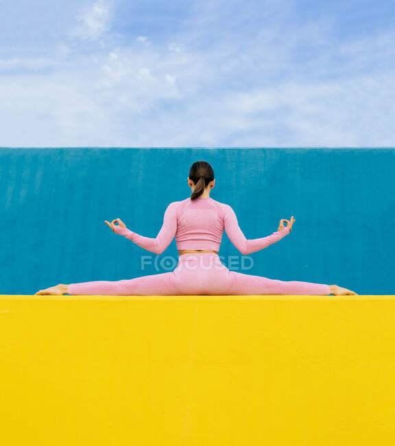 Rückansicht einer schlanken Frau, die im Split sitzt und Gyan Mudra macht, während sie auf einer hohen gelben Wand auf blauem Hintergrund sitzt — Stockfoto