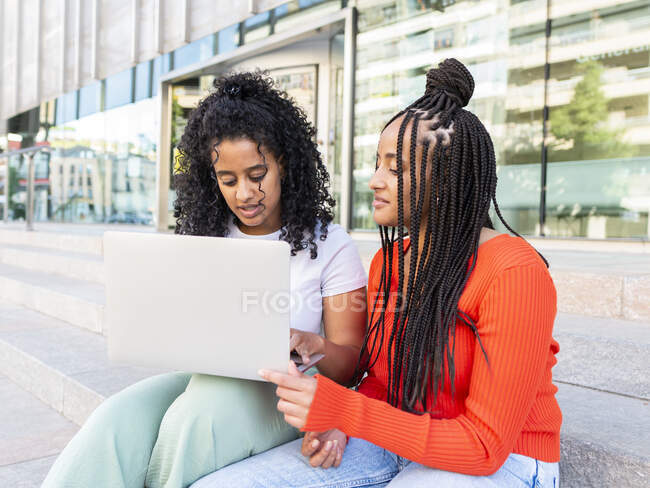 Веселые разнообразные подруги в модной одежде сидят на лестнице и просматривают ноутбук на городской улице в летний день — стоковое фото