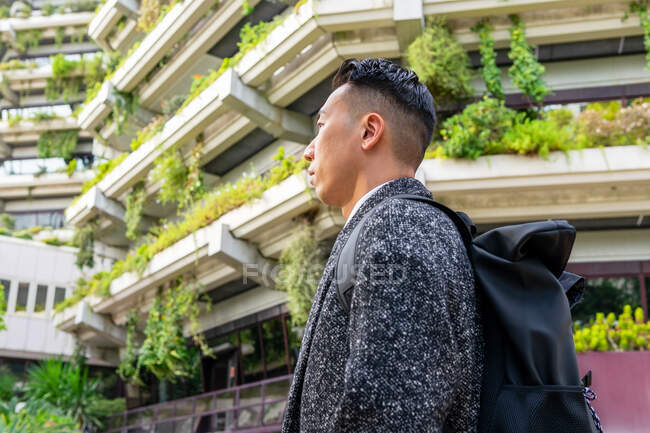 Vista lateral de joven empresario étnico masculino con corte de pelo moderno y mochila mirando hacia otro lado contra el árbol y las casas urbanas - foto de stock