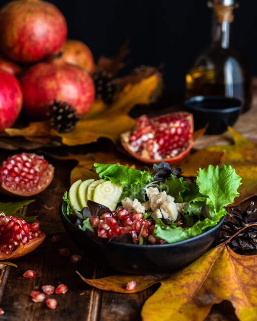 Appetizing pomegranate salad in bowl severed on wooden table with autumn leaves and bottle of olive oil on black background — Stock Photo