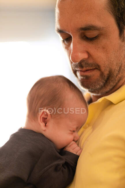 Vista lateral de la cosecha padre adulto en ropa casual abrazando adorable recién nacido suavemente en casa a la luz del día - foto de stock