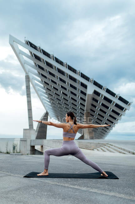 Full body of sportive female in activewear practicing Warrior posture while training on street near solar panel against cloudy sky — Stock Photo