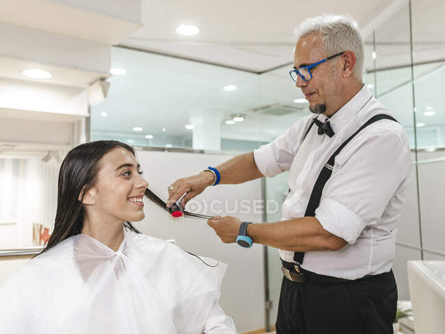 Усміхнений жіночий клієнт у білому мисі дивиться на чоловічу перукарню, що працює з волоссям — стокове фото