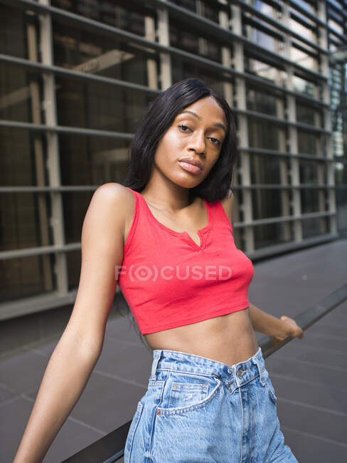 Confident African American female in stylish outfit looking at camera while leaning on railing in city with modern multistory buildings — Stock Photo
