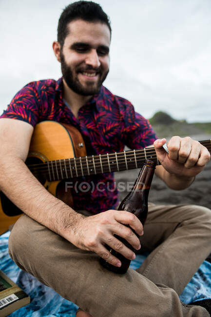 Desde abajo de positivo barbudo músico masculino sentado con guitarra acústica y abriendo botella de cerveza mientras pasa tiempo en la playa en la naturaleza a la luz del día — Stock Photo