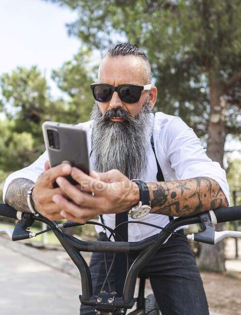 Bearded male in sunglasses and white shirt browsing on smartphone while sitting on bicycle in park with trees — Stock Photo