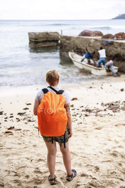 Back view of anonymous traveler in casual clothes and orange rucksack standing on sandy beach and looking at local fishermen in boat — Stock Photo