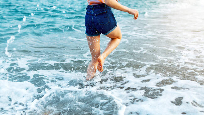 Босиком обрезанная неузнаваемая женщина-путешественница, бегущая вдоль песчаного пляжа, омытая пенными волнами в ветреную погоду — стоковое фото