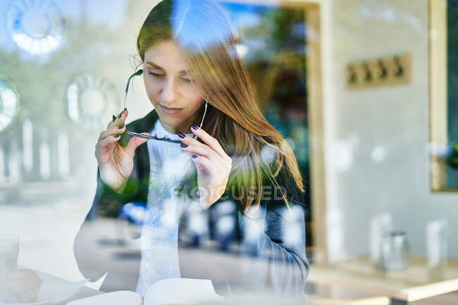 A través de un vaso de trabajadora concentrada quitándose los anteojos mientras lee notas en bloc de notas en la mesa con café y croissant en la cafetería - foto de stock