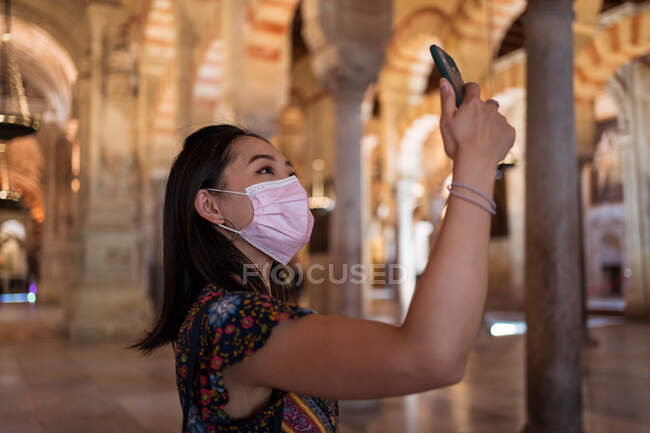 Vista lateral de la viajera asiática en máscara protectora tomando fotos en un teléfono inteligente mientras está de pie en la antigua mezquita durante la pandemia - foto de stock