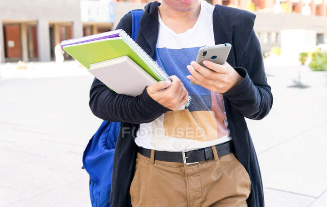 Anonymous male student in casual wear with copybooks text messaging on modern cellphone while standing near university building during studies — Stock Photo