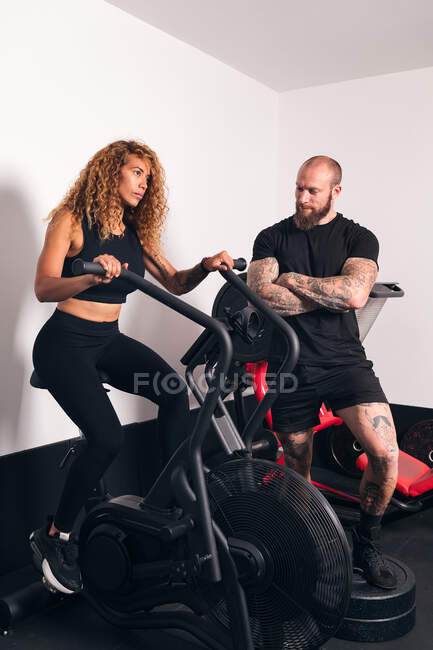 Sportive concentrée avec de longs cheveux bouclés assis sur une machine à vélo et faisant de l'entraînement cardio avec un entraîneur personnel dans la salle de gym — Photo de stock
