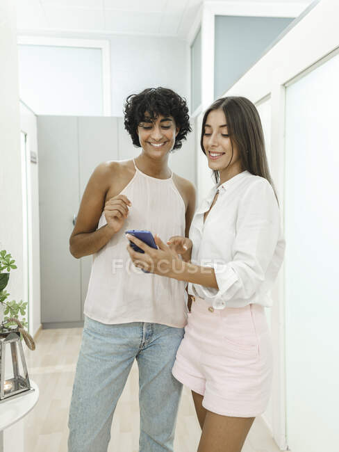 Щасливі молоді багаторасові подружки в стильному одязі, що стоять у світлій квартирі, використовуючи мобільний телефон і дивлячись на екран разом — стокове фото