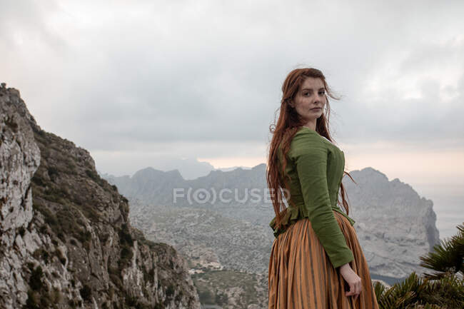 Полный вид на тело мечтательной женщины в старомодной одежде, стоящей на краю каменистой скалы возле моря в туманную погоду — стоковое фото