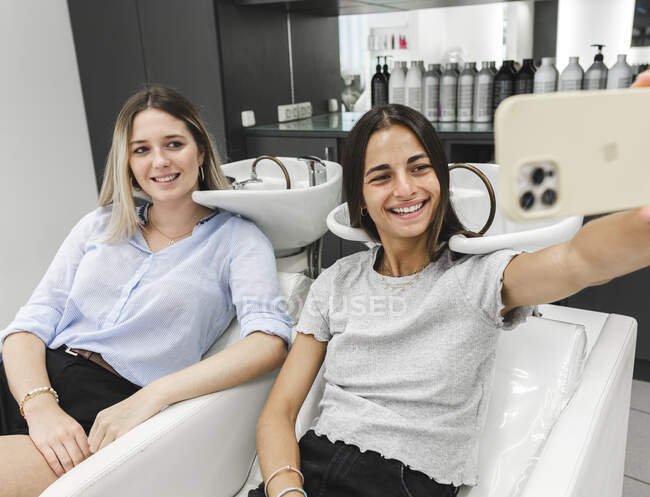 Delightful young woman and friend smiling while sitting in chairs in hairdressing salon and taking selfie — Stock Photo