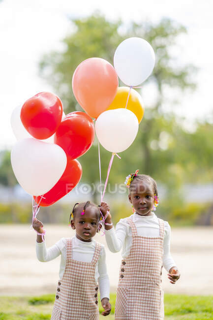Щасливі афроамериканські маленькі сестри в схожих сукнях стоять з барвистими кульками в руках на зеленій траві в парку вдень. — стокове фото