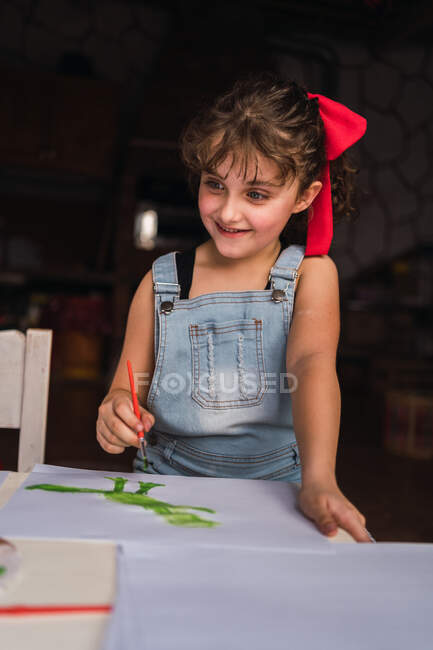 Positive girl in denim overall with paintbrush painting with green paint on white paper while standing at table with chair — Stock Photo