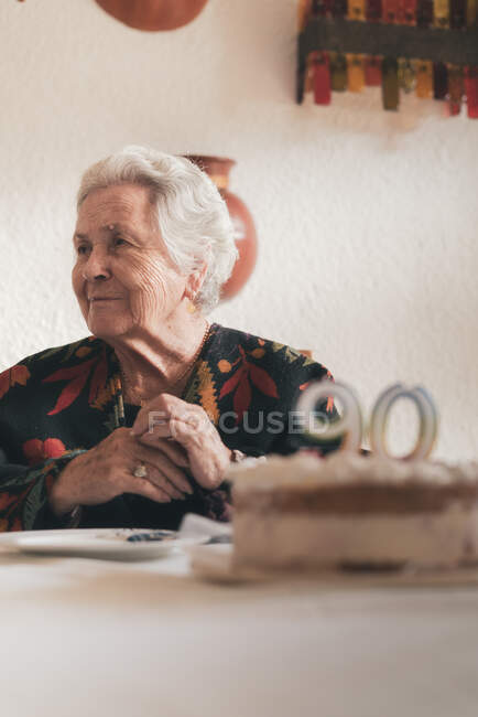 Aged woman blowing candles on birthday cake then clapping hands while celebrating 90th anniversary with relative at home — Stock Photo
