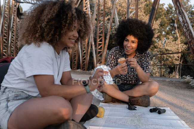 Felici amiche afroamericane che mangiano cibo seduti con le gambe incrociate sul ponte vicino alla mappa e il binocolo durante la pausa in escursione — Foto stock