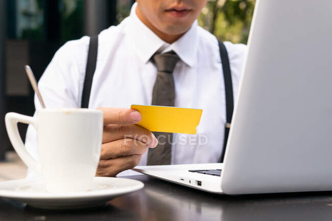 Imprenditore maschio irriconoscibile ritagliato con carta di credito seduto al tavolo della caffetteria urbana con netbook e tazza di caffè durante lo shopping online — Foto stock