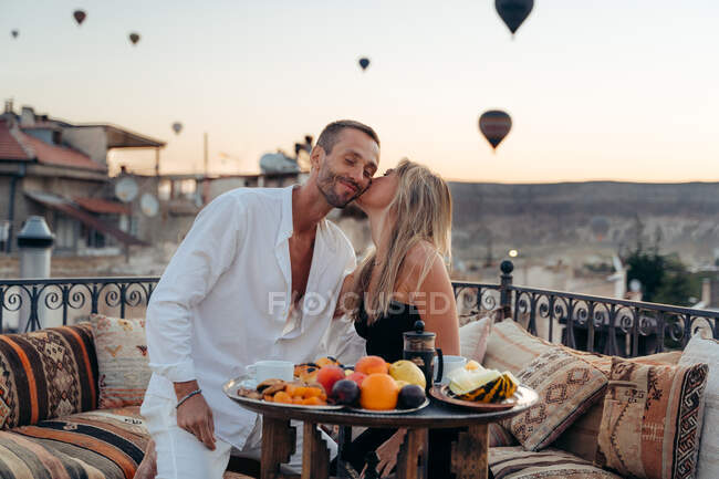 Woman kissing soulmate while sitting in cafe on rooftop terrace and enjoying summer evening on background of hot air balloons — Stock Photo