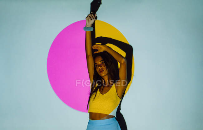 Trendy giovane donna ispanica con braccio sollevato guardando lontano contro l'ombra e luci proiettore colorato su sfondo grigio — Foto stock