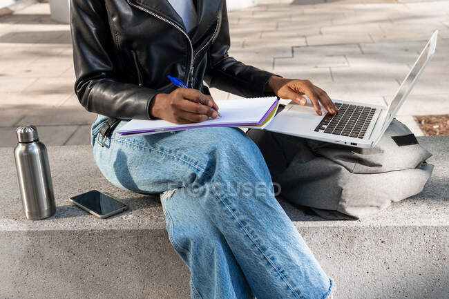 Anonyme Afroamerikanerin in Lederjacke blättert im Netbook, während sie Notizen in einem Notizbuch auf der Straße in der Nähe eines Gebäudes in der Stadt macht — Stockfoto