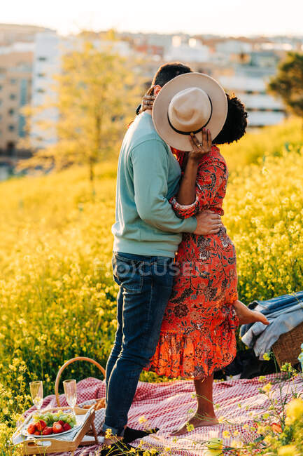 Side view of couple kissing and embracing while covering face with hat in blooming meadow in sunny day — Stock Photo