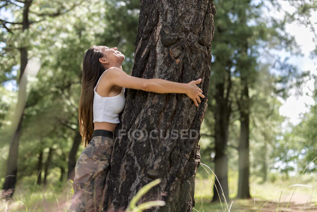 Mujer tranquila en ropa casual con los ojos cerrados abrazando tronco de árbol grueso en el bosque con hierba verde borrosa durante el senderismo - foto de stock