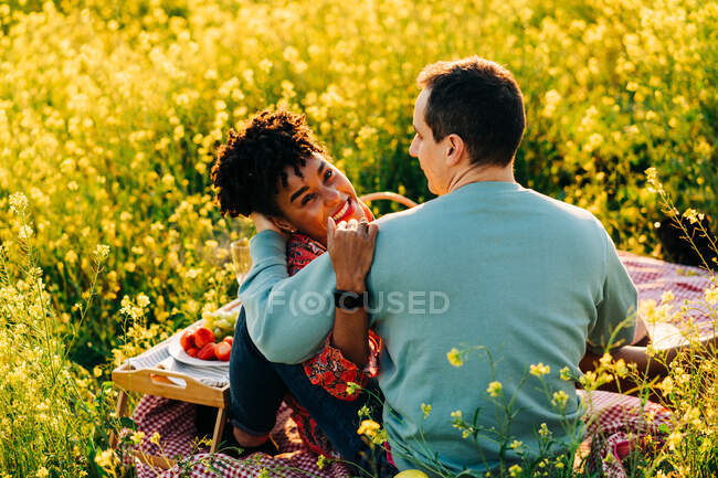 Jovem senhora afro-americana com cabelo encaracolado abraçando namorado com olhos fechados durante piquenique no prado florido no dia ensolarado — Fotografia de Stock