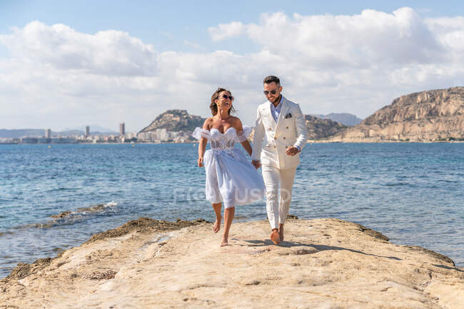 Full body of cheerful barefoot wedding couple running on shore near rippling sea while enjoying wedding day in sunny nature — Stock Photo
