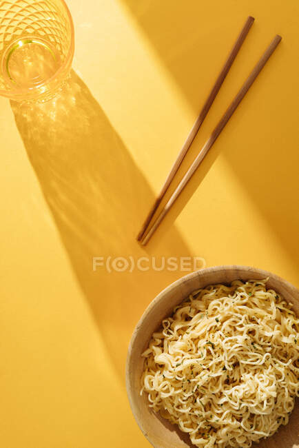 Bol de délicieuses nouilles avec assaisonnement placé sur fond jaune avec baguettes en bois et verre dans la pièce lumineuse — Photo de stock