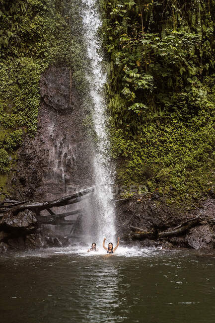Des voyageuses méconnaissables nageant dans un étang sous la puissante cascade de Sao Nicolau qui coule à travers une falaise rocheuse couverte d'une végétation verte luxuriante — Photo de stock