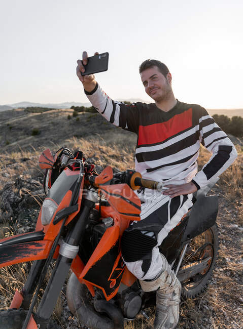 Contenuto motociclista maschile in costume sportivo scattare autoritratto su smartphone mentre seduto sulla moto moderna durante le corse in natura — Foto stock