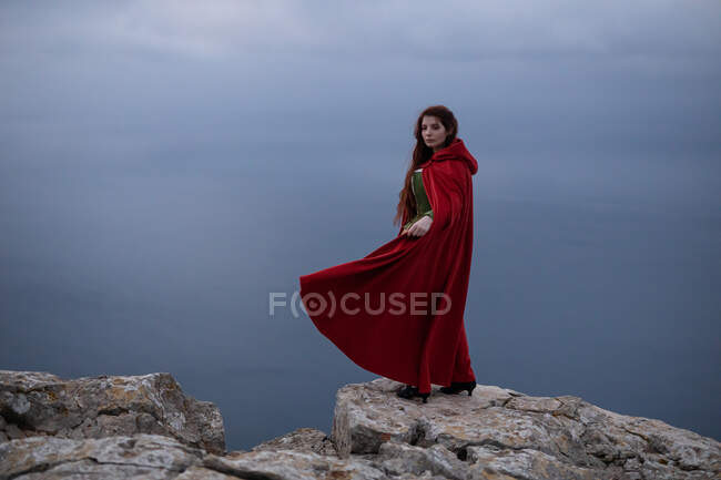 Мрійлива жінка з довгим волоссям у вікторіанському вбранні з плащем над безконечним морем проти хмарного неба в природі. — стокове фото