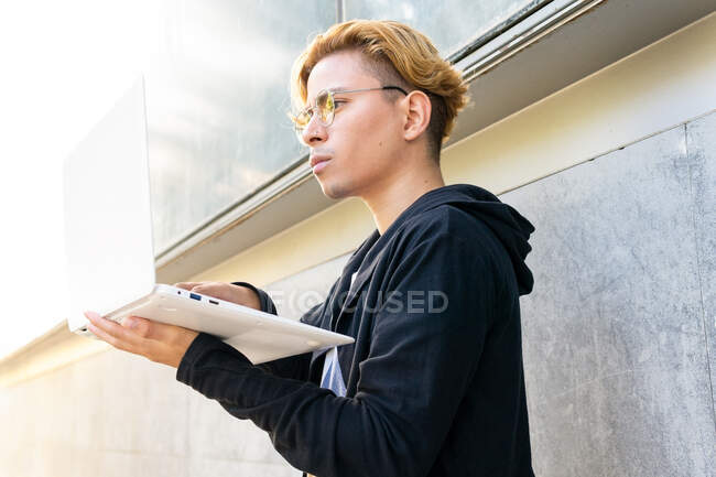 Freelancer masculino joven concentrado escribiendo en netbook moderno mientras está parado en la calle en la ciudad durante el trabajo en línea - foto de stock