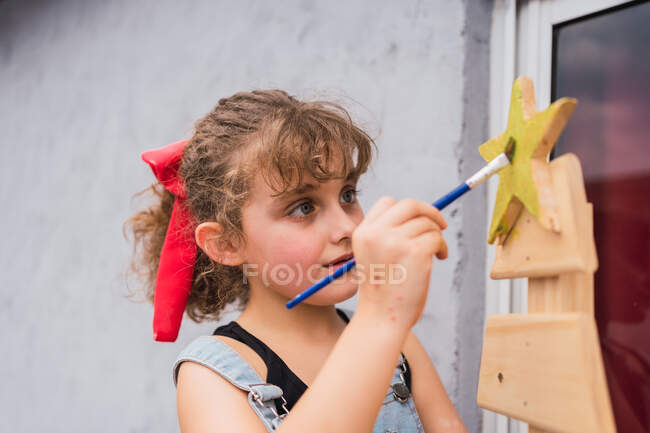 Focused girl in denim overall painting with paintbrush on decorated Christmas tree in light room during holiday preparation at home — Stock Photo