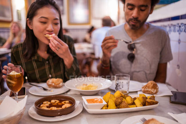 Врожай голодний багаторасовий пара з темним волоссям в повсякденному одязі, що їсть апетитні страви в сучасному ресторані під час обіду — стокове фото