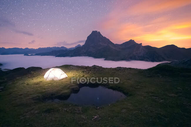 Светящаяся палатка для кемпинга, расположенная на травянистой местности против горного хребта в природе Испании с густым туманом в вечернее время — стоковое фото