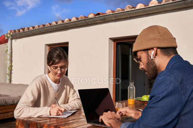 Mujer concentrada en anteojos escribiendo en planificador en la mesa con compañero de piso masculino en auriculares inalámbricos escribiendo en el portátil en el balcón - foto de stock