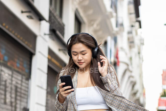 Ethnische Frau mit langen braunen Haaren checkt Handy und hört Lieder in drahtlosen Kopfhörern in Madrid — Stockfoto